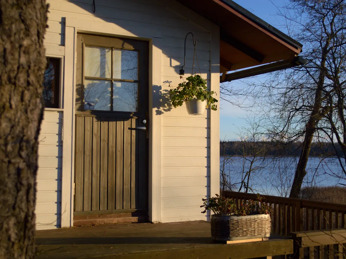 Vaalean mökin ovi ja kuisti, taustalla järvi koivujen välistä.