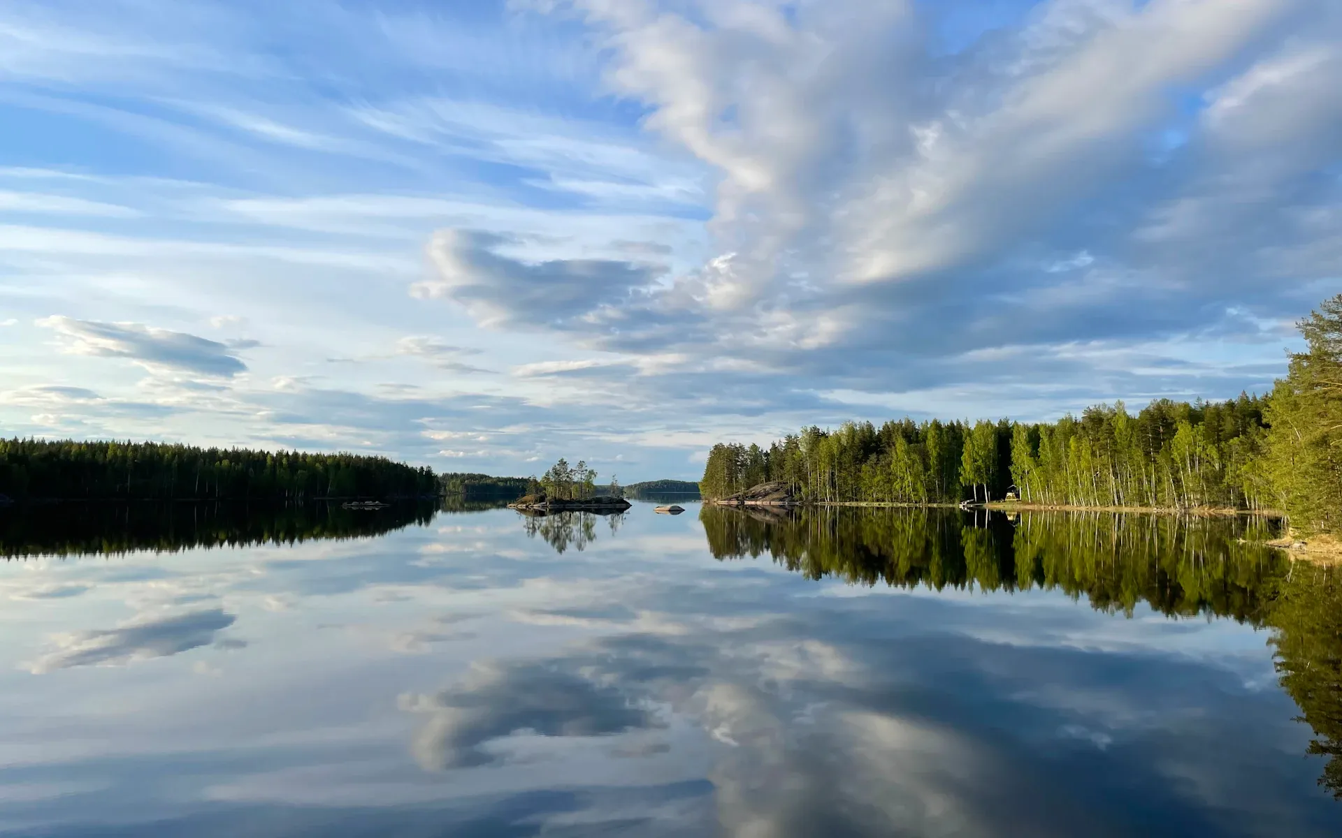Tyyni järvi heijastaa pilviä, rannoilla havumetsää.