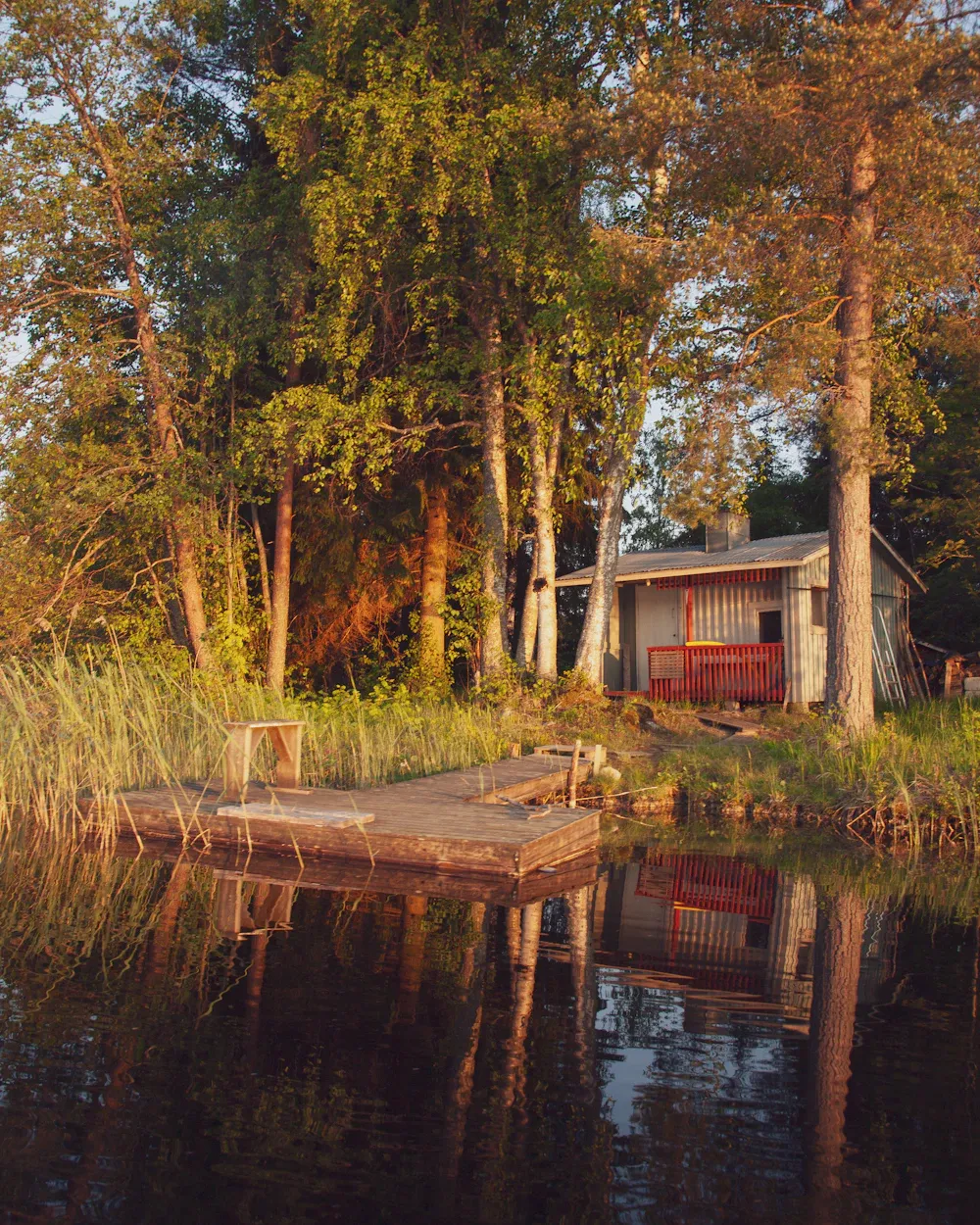 Punainen rantasauna ja laituri ilta-auringossa, puut heijastuvat veteen.