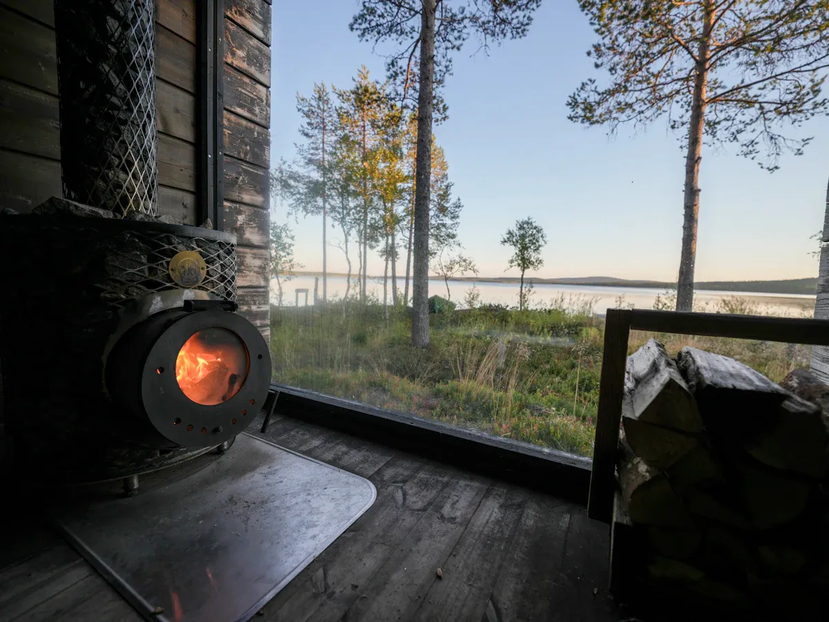 Kamiinassa palaa tuli, ikkunasta näkyy mäntyjä ja järvi.